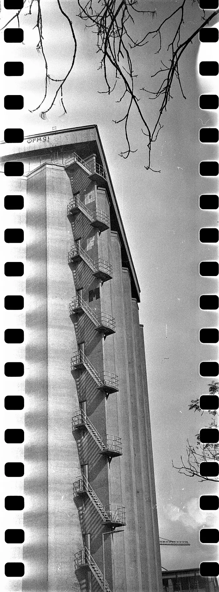 Vertical panorama of a tall industrial silo with a fire escape stairway running criss cross down the side. It shows dust marks and streaks from faults in the development process.