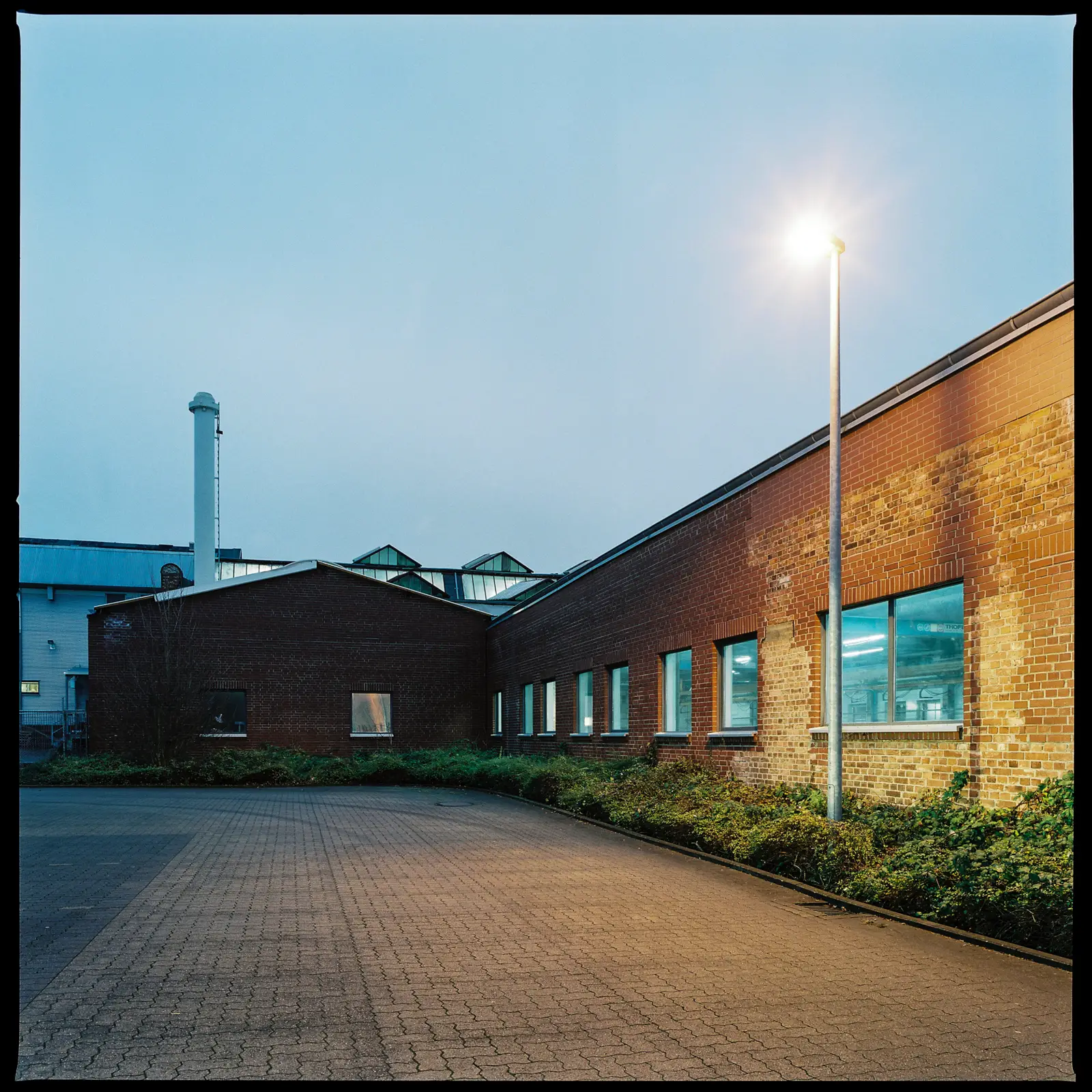 6x6 photograph of a run-down factory building at dusk, with parking spaces in the foreground