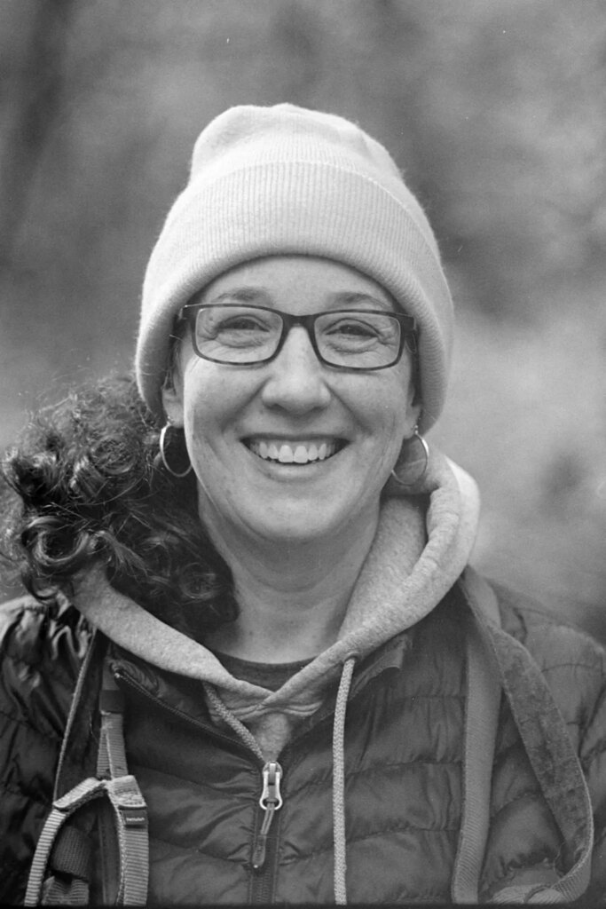 A black and white photo of a woman wearing a sweatshirt, knitted hat and glasses.