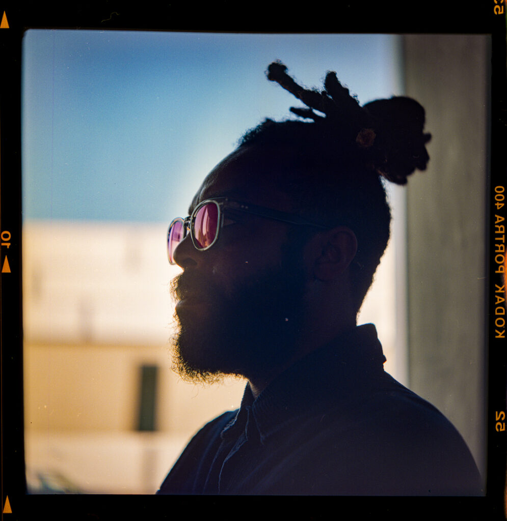 A black man in partial silhouette with his purple sunglasses reflecting their color.
