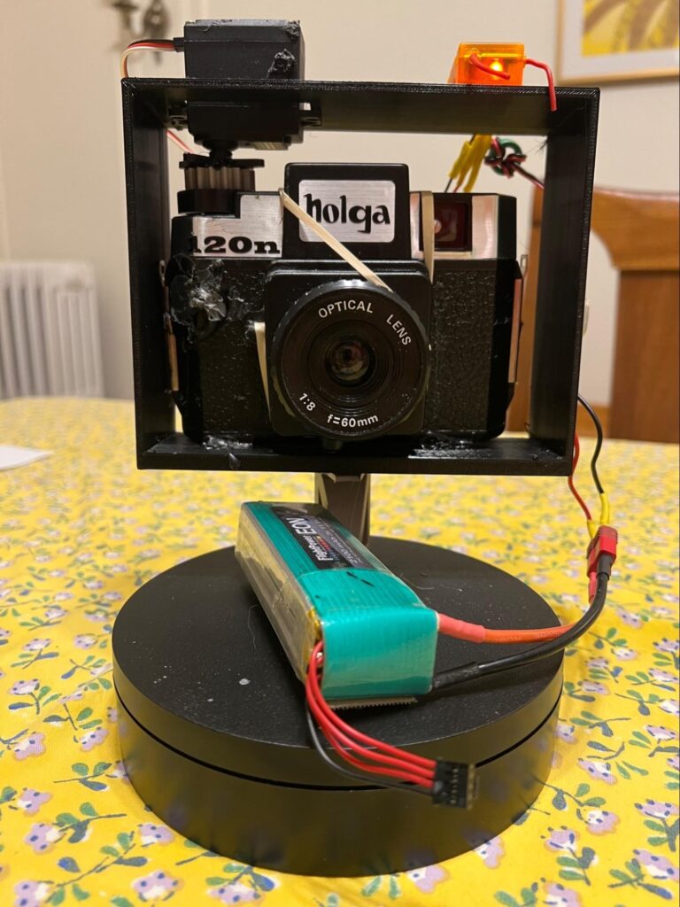 A photo of a holga camera sitting in a square plastic frame that is lifted off the electric turntable by a short plastic extension. There is a battery sitting on the turntable. 