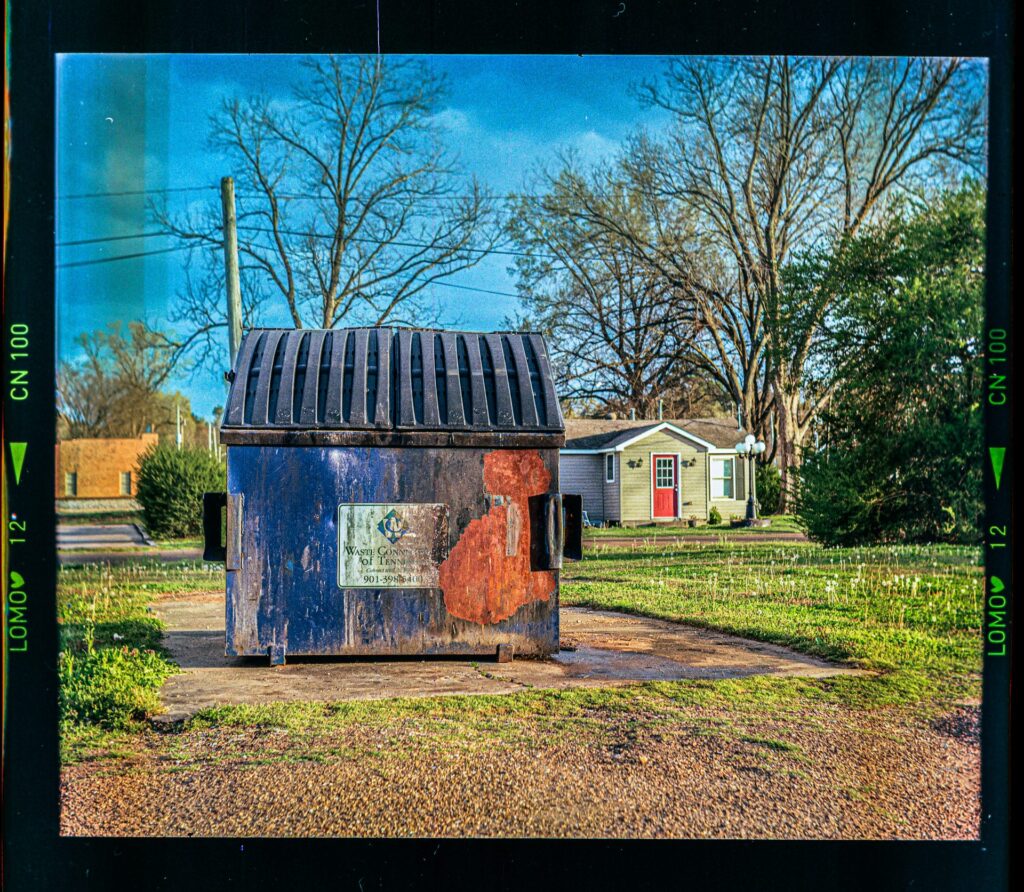 A dirty and rusted dumpster stationed on a cement square with a house in the background