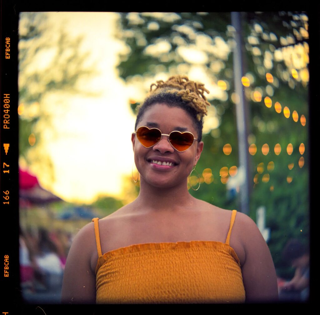 An light skinned african american woman with short blonde locks, wearing heart shaped sunglasses and smiling.