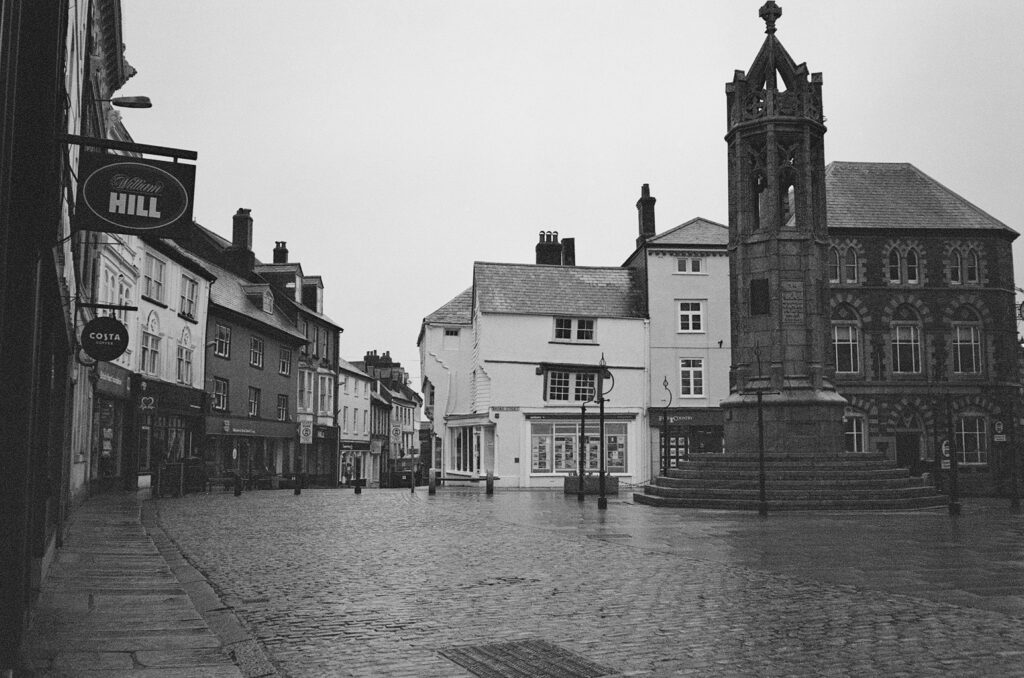 Black and white photo of cobbled town square with a variety of old buildings surrounding it and a large war memorial in the middle.