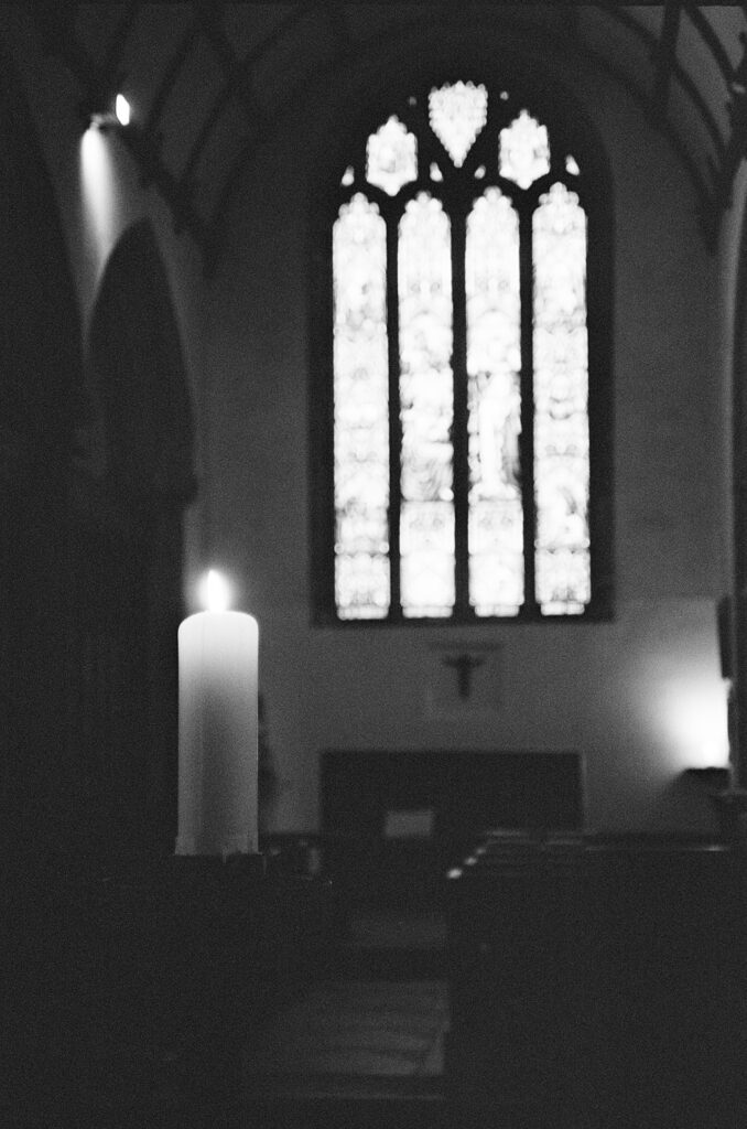 Black and white photo of a church interior, with a lit candle in the foreground and an out of focus stained glass window in the background