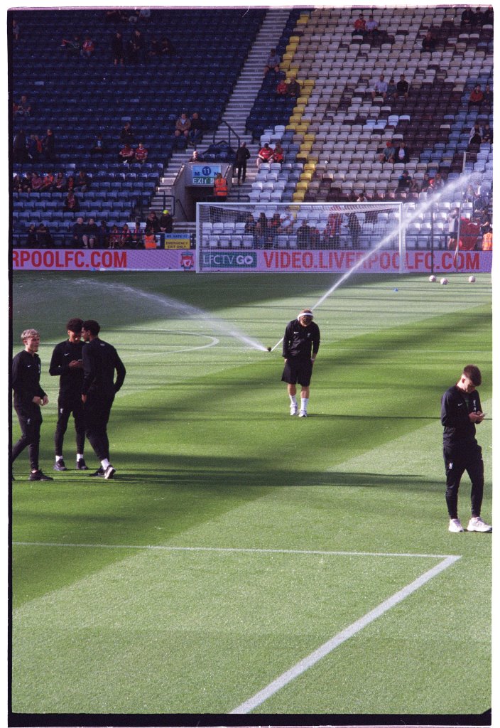 Harvey Elliot stands on the pitch at Deepdale in headphones. To the right we can see Bobby Clark, Calum Scanlon and James McConnell; to the right Ben Doak