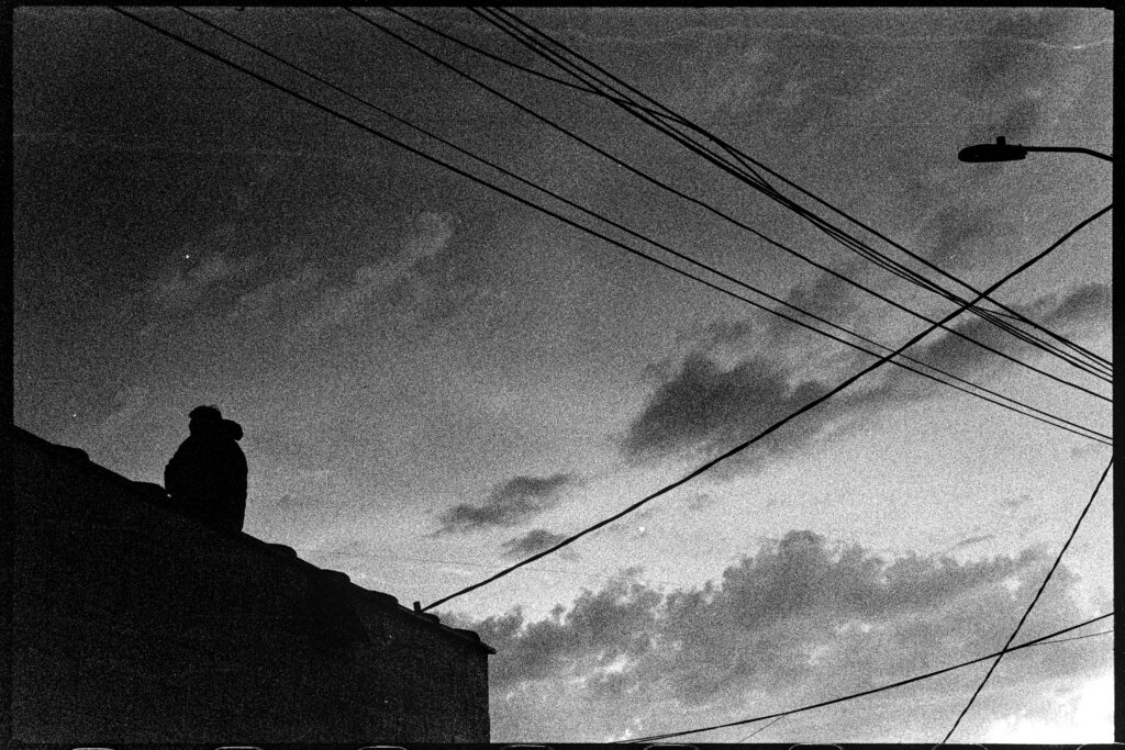 A man, sweeping the roof of his building, pauses to watch the sky to the west.
