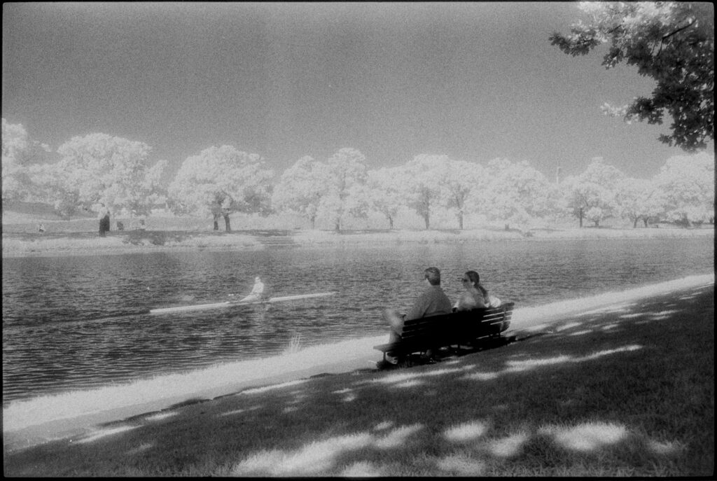 Black and white infrared image of a river scene, two people on a bench seat watch as a rower makes his way down the nearby river. Trees in the background