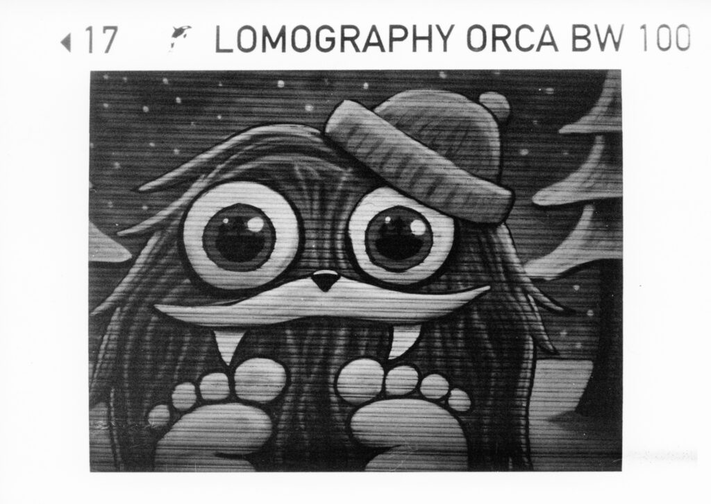 Black and white photo of graffiti art, a cute hairy creature with big eyes and fangs wearing a beaning