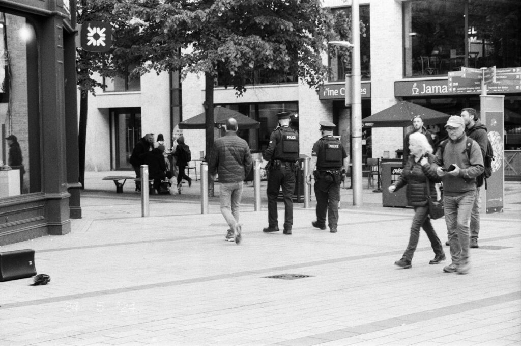 Two PSNI officers on patrol in Belfast city centre with other civilians/pedestrians in frame.
