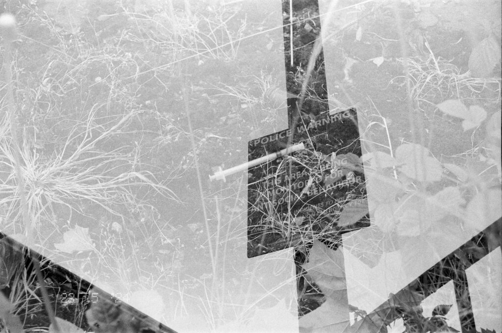 Black and white image. Double exposure on Ilford HP5 film showing a discarded hypodermic needle with a police sign warning against anti-social behavior
