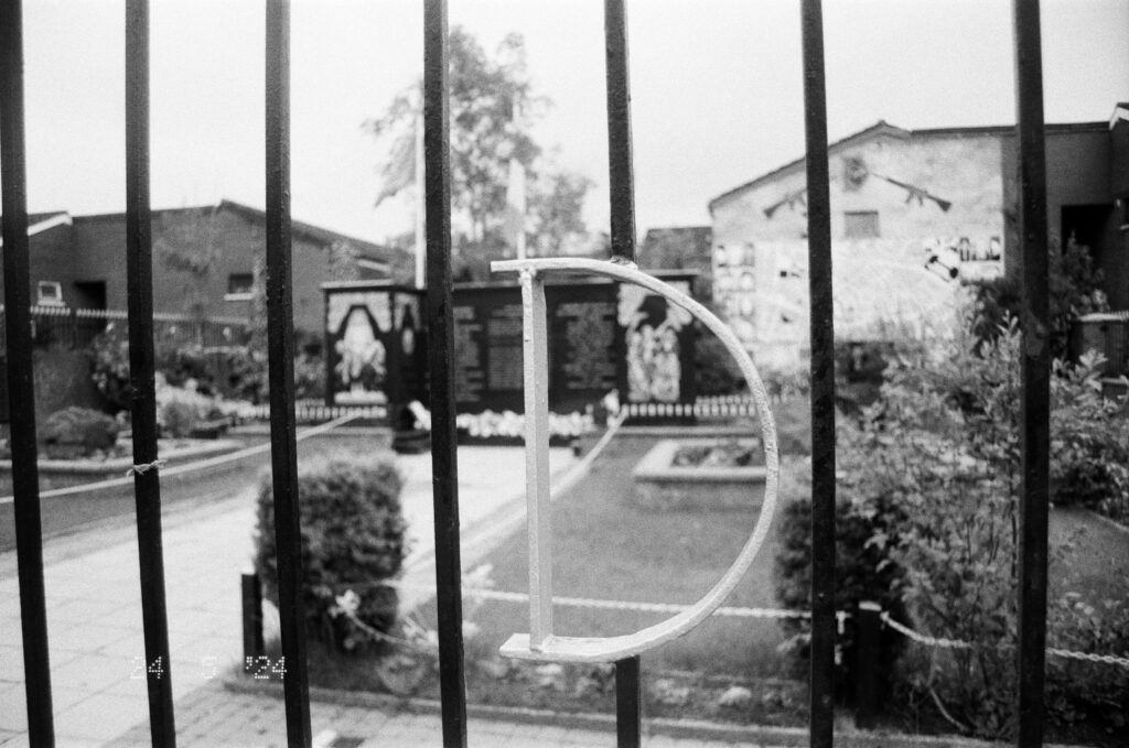 A picture of the 'D Company' PIRA memorial garden in West Belfast. The image focuses on the letter D in the wrought iron fence enclosing the garden, while the garden itself is in soft focus in the background.