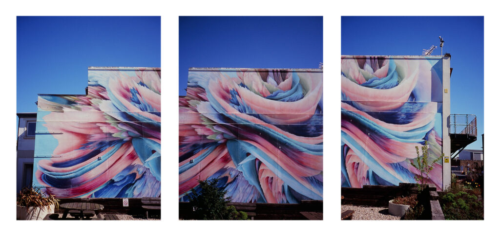 A colour triptych image, three portrait photos of a mural on the side of a building. The mural is abstract with big sweeping waves of rainbow ribbons in a pastel colour.