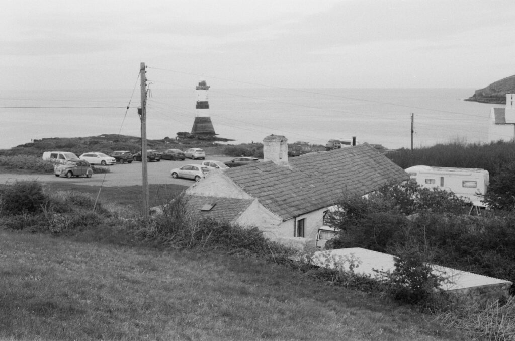 A general image of Penmon Point with the sea in the distance, a lighthouse, parked cars with a white building in the foreground.
