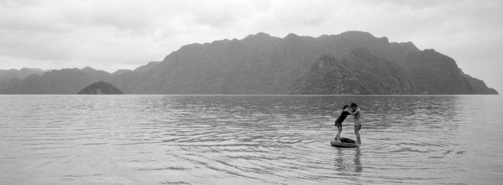 Two kids playing on an inflatable tire in the water with a stunning backdrop of islands in the distance in black and white
