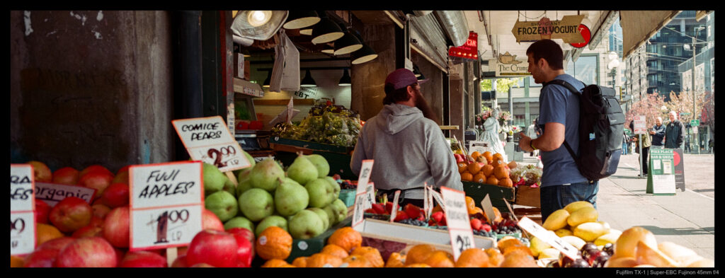 A worker and shopper discuss produce at a colorful streetfront display.