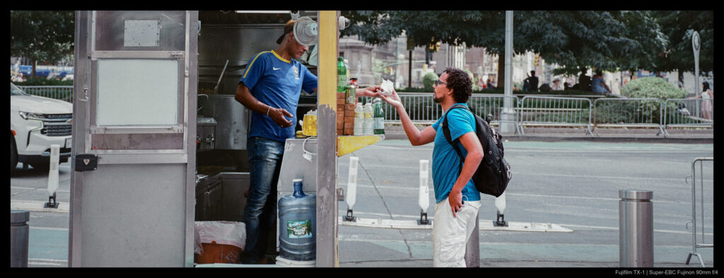 A view of a sale happening at a food cart viewed from the side such that the owner and customer are on opposite sides of the cart front.