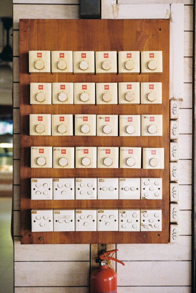 Close-up shot of a worn wooden panel mounted on a tiled wall, displaying four rows of vintage, cream-colored rotary fan speed controllers and two bottom rows of electrical sockets. A small red fire extinguisher is mounted directly beneath the panel.