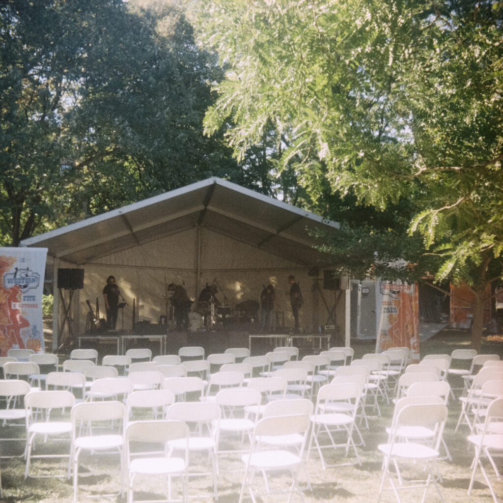 A band warming up on a small scene. Several rows of unoccupied white chairs are lined-up.