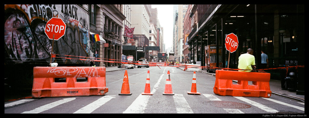 A person sits on a safety barrier arranged across a street with some traffic cones, their back to the camera and showing a high-vis vest.