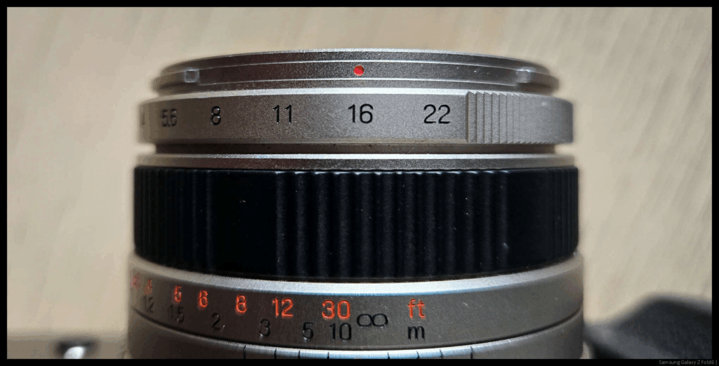 A top-down image of a Fujifilm TX-1 lens with the aperture ring set to f/16.