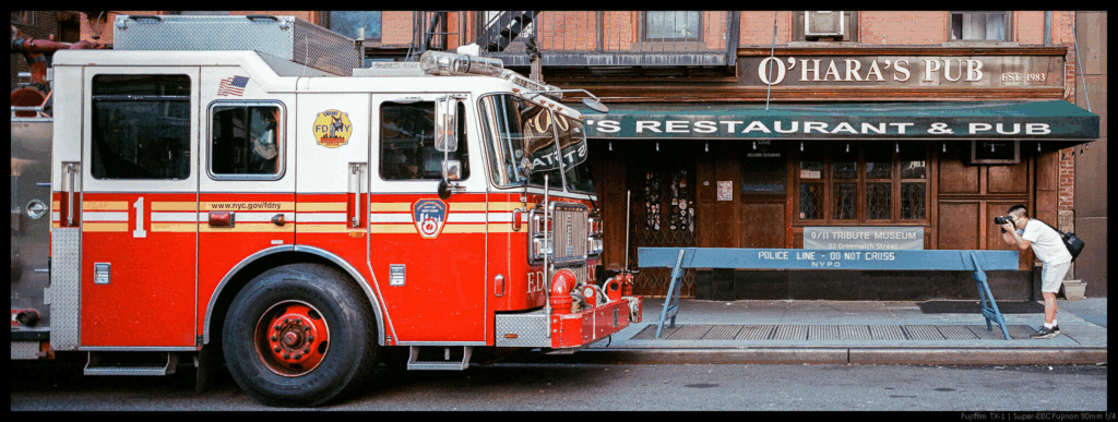 An image of a person photographing a FDNY fire truck, with a police barrier between them endwise.