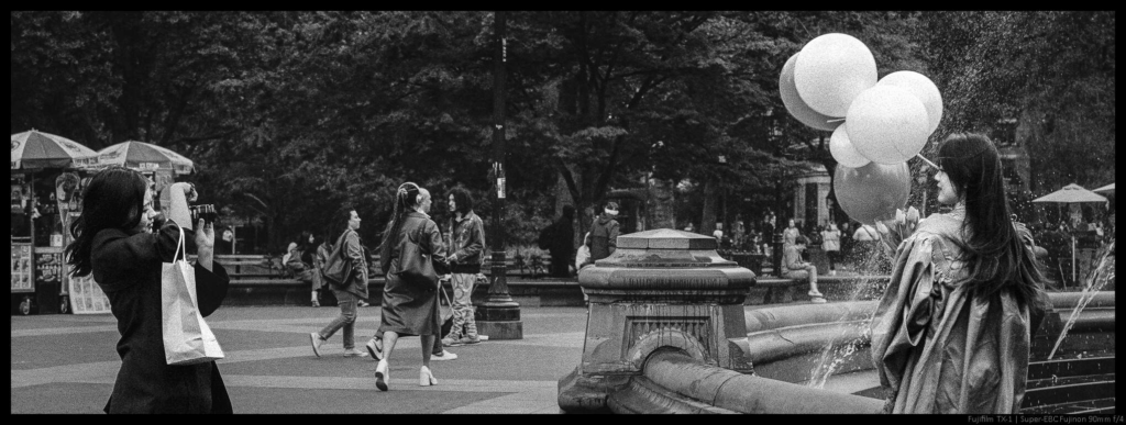 An image of a person taking a photo of their friend dressed in a graduation cap and gown while holding balloons.