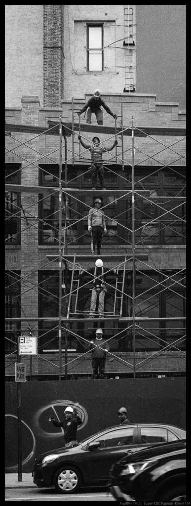 A number of construction workers stand on different ascending levels of scaffolding, passing large metal frames up the line.