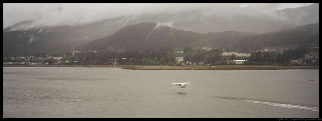 An image of a plane taking off, trailing a small foam wake, from a small inlet. Across the water are a series of low mountains and a small number of buildings.