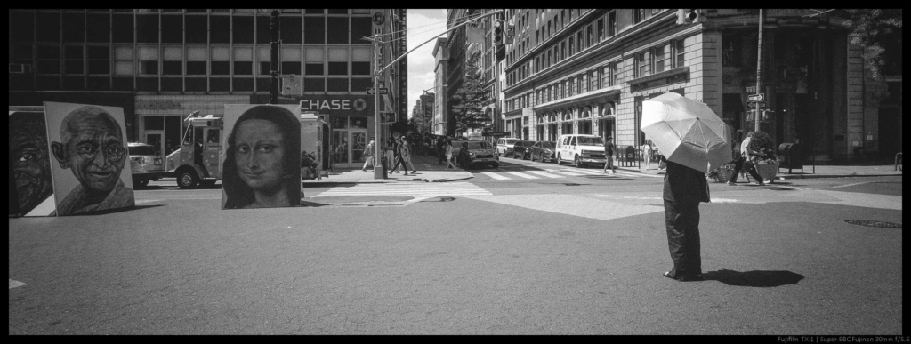 A photo of a person standing beneath an umbrella looking to the left at two paintings (a large copy of the Mona Lisa and a large image of Mahatma Gandhi). The eyes of the paintings appear to be looking back at the person.
