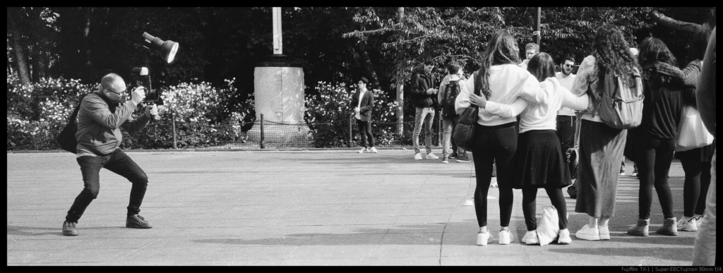 An image of a photographer crouching down to line up a shot with a large camera and flash setup of a group in Washington Square Park.