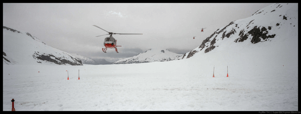 An image of three helicopters coming in for landing on a white glacier in the mountains.