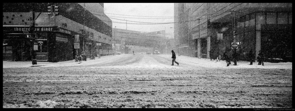 An image of two people crossing a snow-strewn street during a heavy snowstorm between two large sets of buildings.