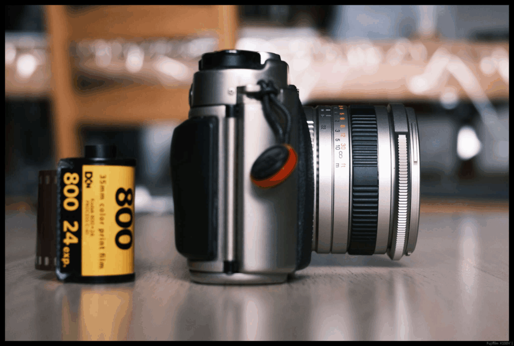 An image of the Fujifilm TX-1 with the 30/5.6 mounted from the side. A 35mm cassette is to the left.