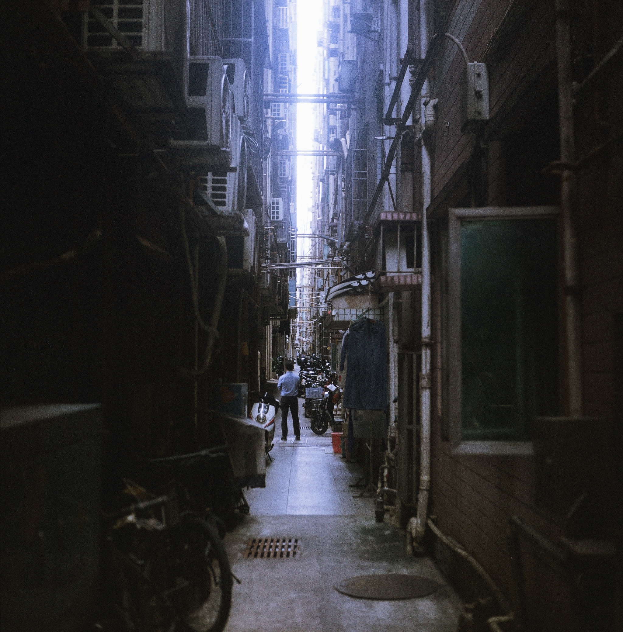 A figure stands alone in an alley between massive buildings on either side.