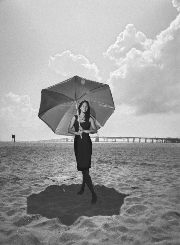 An actor with an open beach umbrella on their shoulders smiles for the camera.