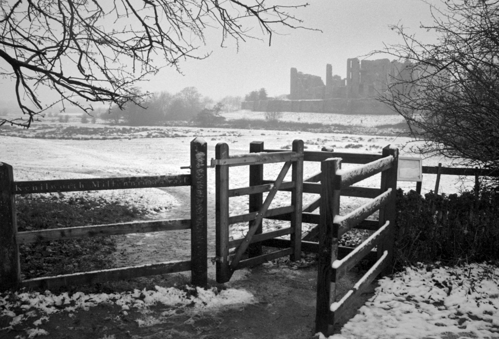 Black and white film photo of a snowy field with misty castle beyond, seen through a foreground kissing gate.