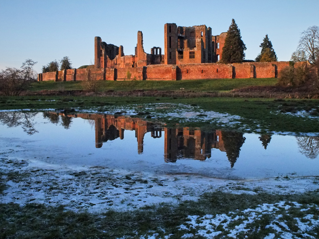 Colour film photo of Kenilworth Castle, see reflected in a flood caused by thawing of the snow and ice seenin the foreground. The castle is glowing in the afternoon sun. The sky is pale blue.