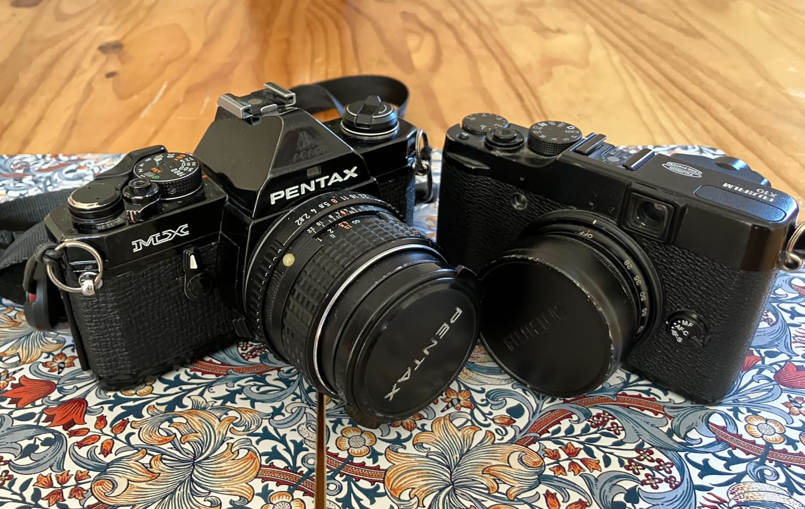Two cameras plaecd close together on decorative mats on a table. The larger is my most ised camera, a Pentax MX with a Pentax 35mm lens, the smaller is my only digital camera, the lovely little Fuji X10 with its cracking zoom lens.