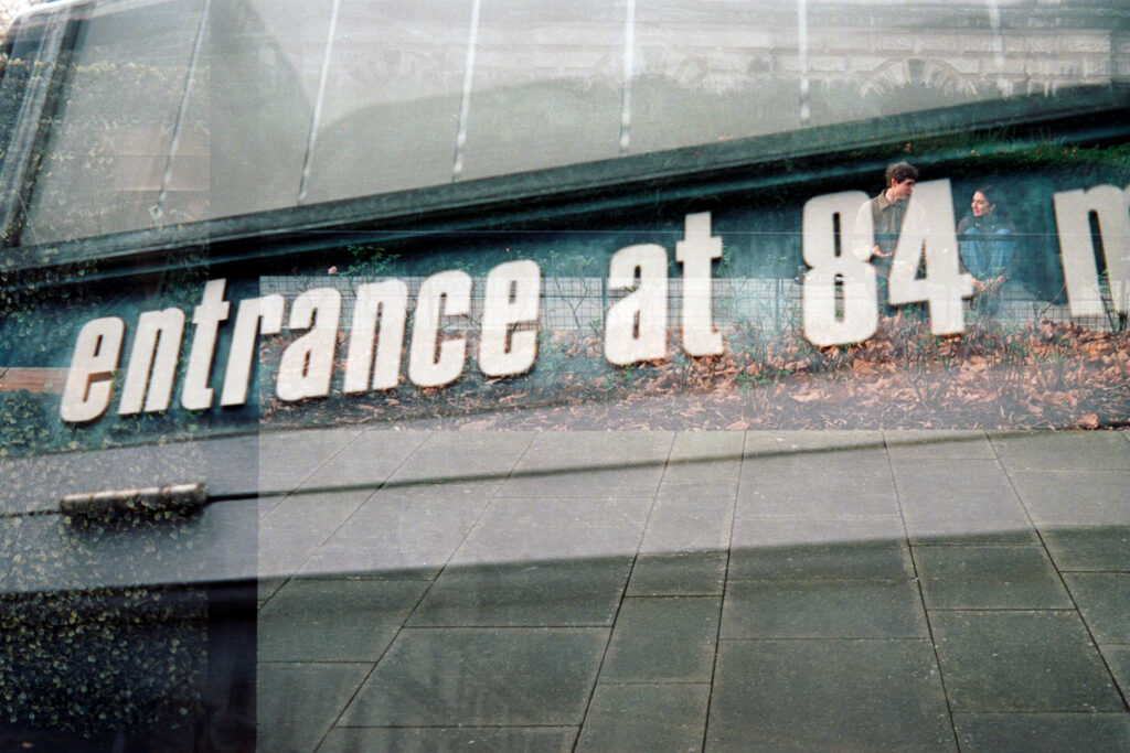 Double exposure on 35mm film featuring a building sign entrance at 84 with a couple sitting in a garden talking. 