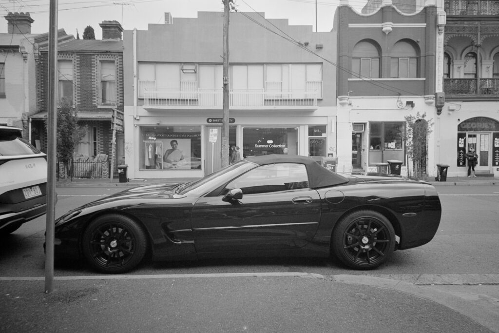 Black and white photo of a Corvette Car parked on the street. Photos is of the side profile of the car. In the background are shops on the other side of the road.