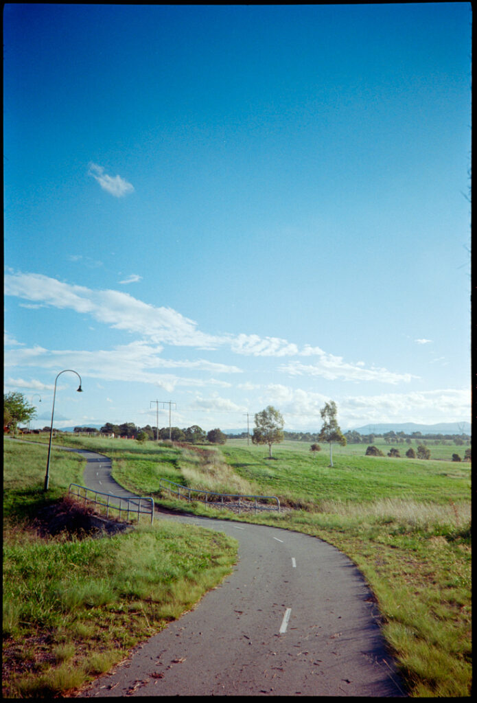 Colour photo of a bike path winding its way down a small slope into a hollow and back up the other side. There are a couple of small trees off to the side in the distance. The sky has a gradient from light blue to dark blue. There are some white clouds on the horizon.