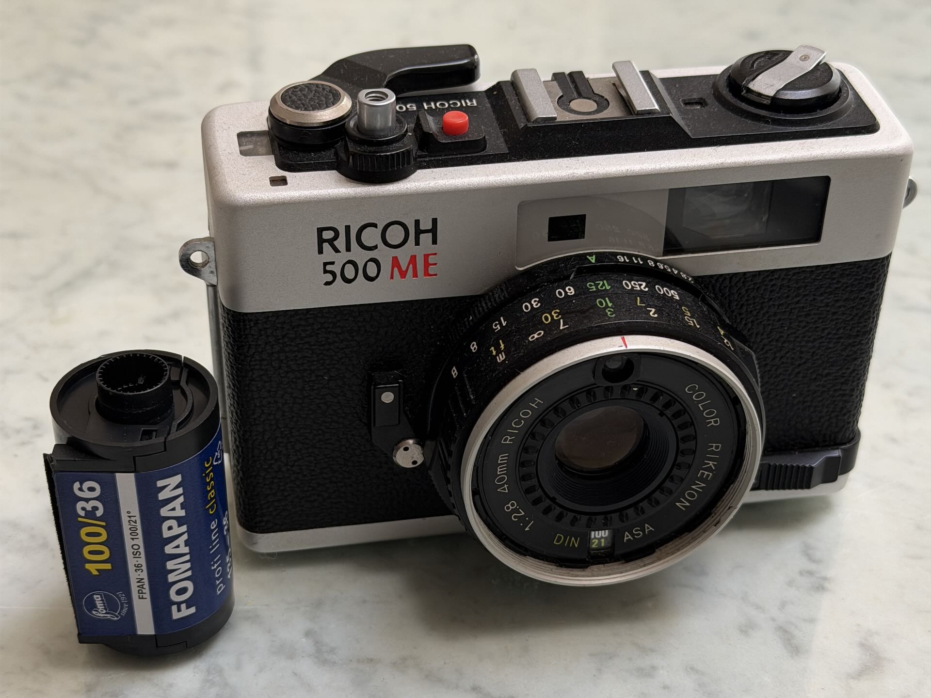 Ricoh 500 ME film camera with 40mm lens next to a roll of Fomapan 100 black-and-white film.