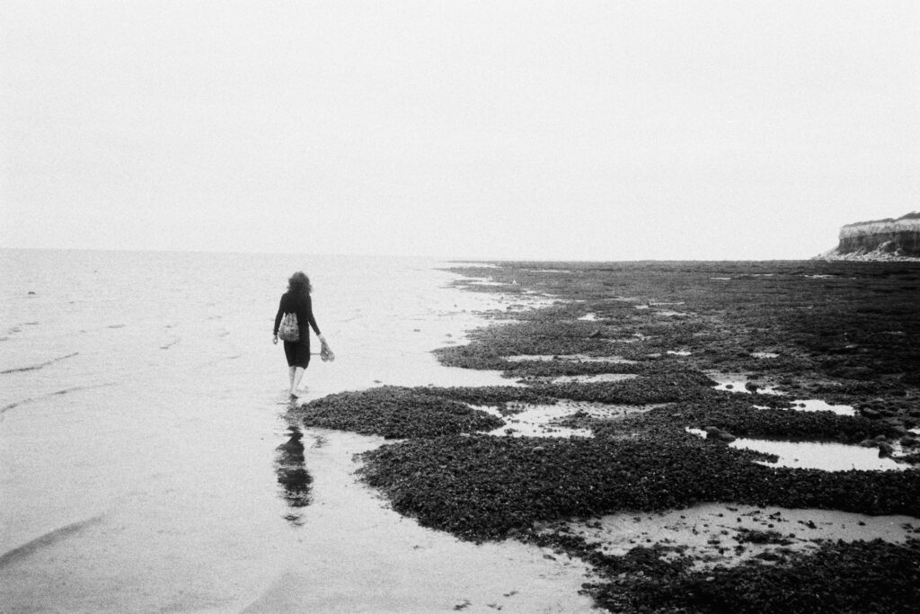 Black-and-white image of a woman walking along the shoreline between water and rocks, with her reflection in the water
