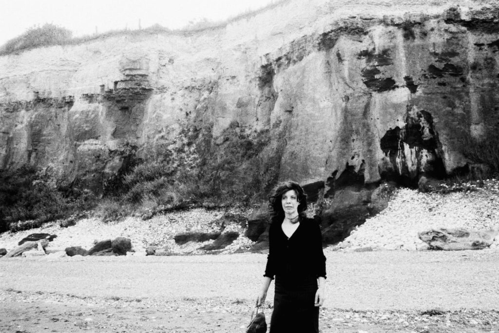 Black-and-white image of a woman standing on a beach in front of textured cliffs, with visible grain softening the background.