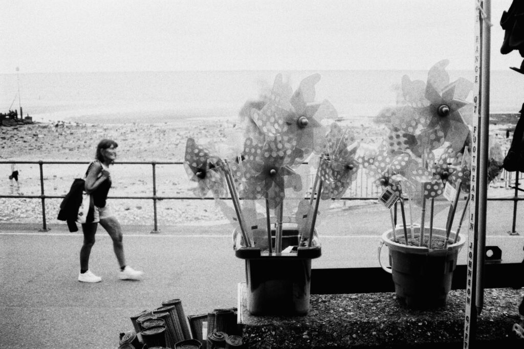Black-and-white image of pinwheels in buckets by the seaside, with a blurred person walking in the background.