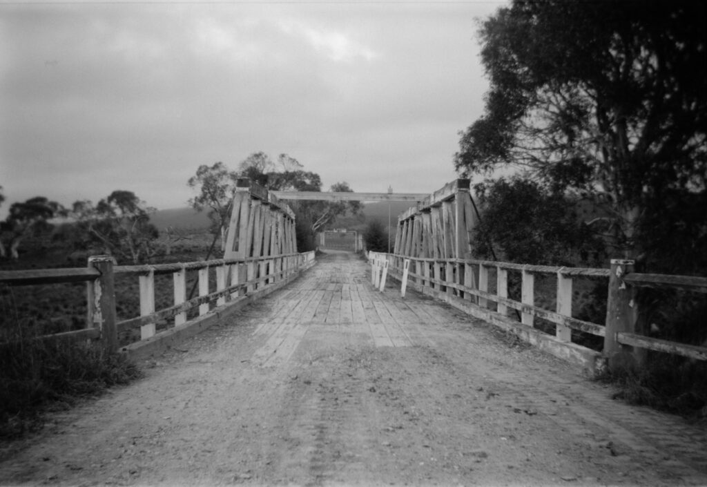 Black and white photo of a wooden bridge on a dirt road. There are some trees on the right side of the image just off the road.