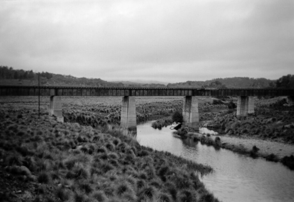 Black and white photo of a railway bridge over a small river. The sky is cloudy. There are small bushes and grasses on the embankments down to the water.