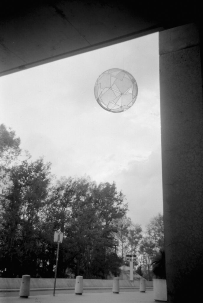 Black and white photo of a sphere suspended above the road outside The National Gallery of Australia. There are bollards in the foreground next to the road and trees in the distance. 