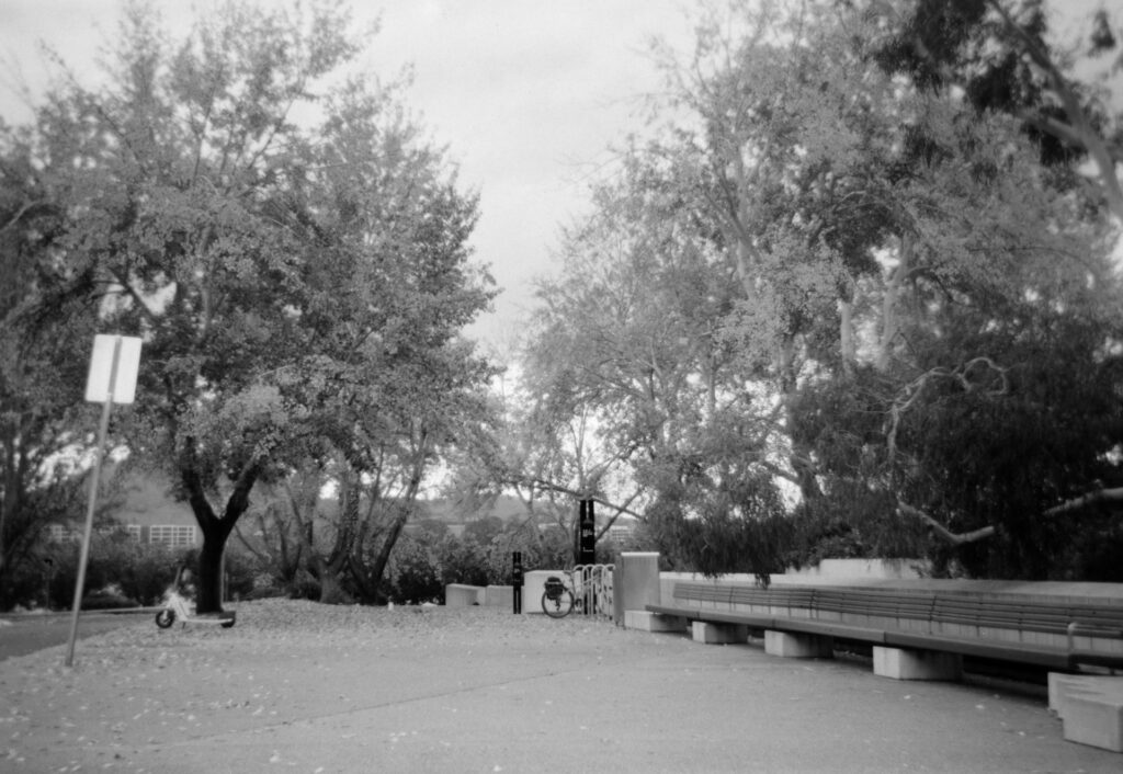 Black and white photo of a wide path with a bench running down the right side and a bike parked at a bike stand at the end of the bench. There are trees in the background and an electric scooter parked on the left.